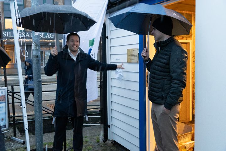 To mennesker holder paraplyer ved en målehytte. En av dem peker på en plakett på veggen.