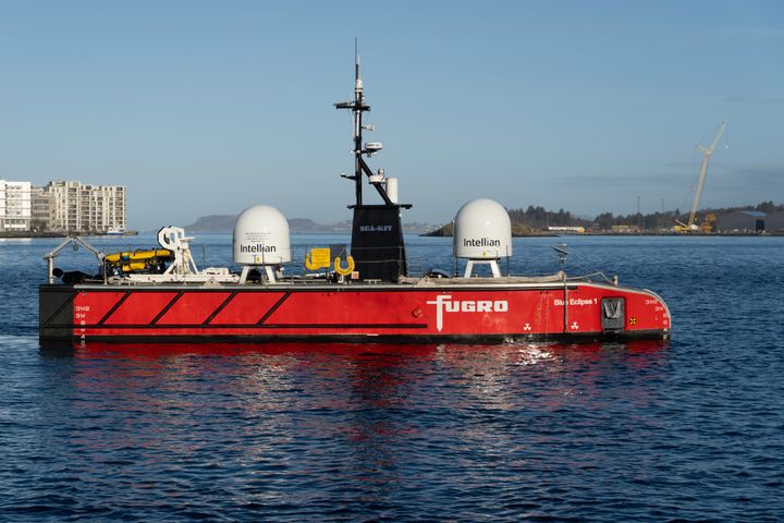 En stor rød USV ved navn Blue Eclipse® fra Fugro, ligger til sjøs med bylandskap i bakgrunnen.