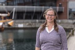 Dame med briller står utendørs ved vann med moderne bygninger i bakgrunnen.