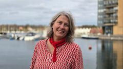 En kvinne i en rød og hvit-mønstrete skjorte står foran en havn med båter.