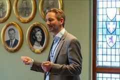 En mann i dress snakker i et rom med flere innrammede portretter på veggen.