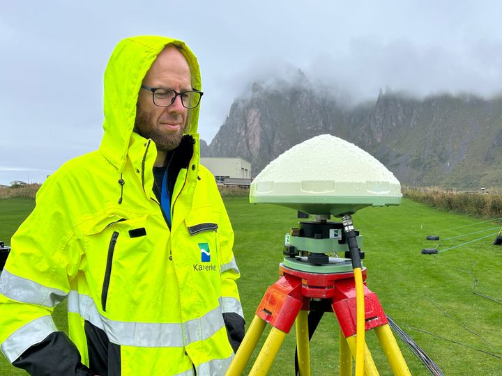 En person i gul jakke står ute ved en GNSS-mottaker i et grønt landskap, med fjell i bakgrunnen.