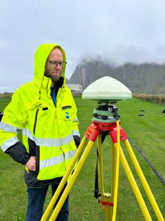En person i gul jakke står ute ved en GPS-stasjon i et grønt landskap, med fjell i bakgrunnen.