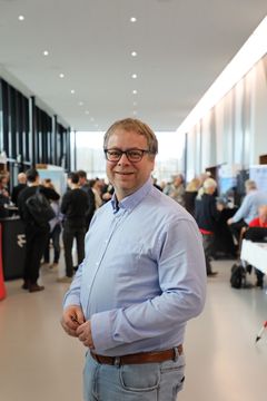 En mann i lyseblå skjorte står og smiler i en moderne, lys innendørs lokasjon med folk i bakgrunnen.