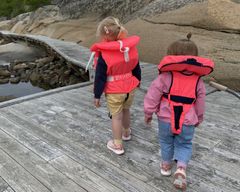 To barn går på en brygge med ryggen mot kamera. De har begge på seg redningsvester. Foto.