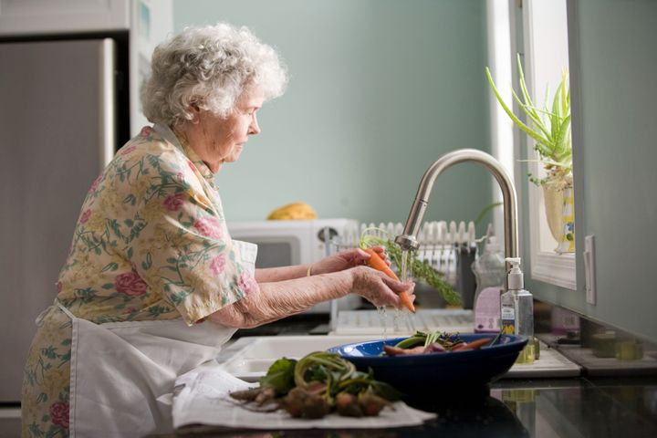 Eldre dame med grått hår står foran en vask og skyller en grønnsak.