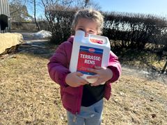 Jente på 3-4- år holder en stor flaske med terrasserens foran segg. I bakgrunnen ser man litt snø og dødt vårgress. Foto.