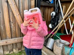 Liten jente med rosa boblejakke holder opp en stor flaske med rosa innhold og et tydelig merke med gift foran ansiktet. I bakgrunnen sees en bod. Foto.