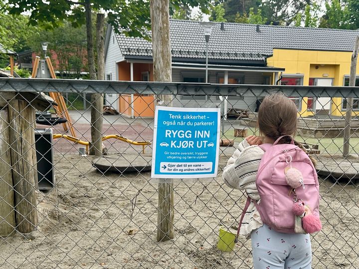 Jente som lener seg mot et gjerde foran en barnehage. Det henger et skilt på gjerde der det står: Rygg inn - kjør ut.