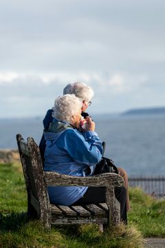 To eldre personer sitter på en benk og ser utover havet.