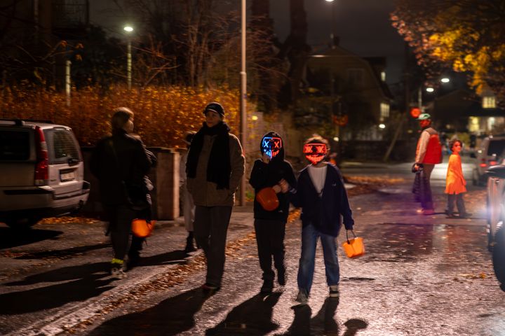 Barn med Halloween-masker og godteribøtter går på en gate om kvelden, med voksne i bakgrunnen.