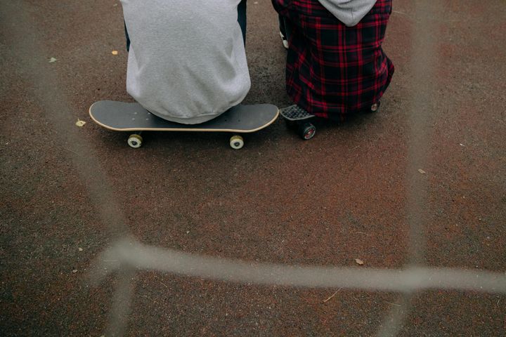 To ungdommer sitter med ryggen til, på hver sin skateboard, på en rød asfaltflate. Bildet er tatt gjennom et gjerde.