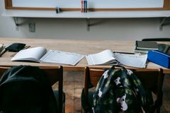 To skolepulter med åpne skrivebøker, arbeidsark og skolesekker hengende på stolene, sett forfra i et klasserom.