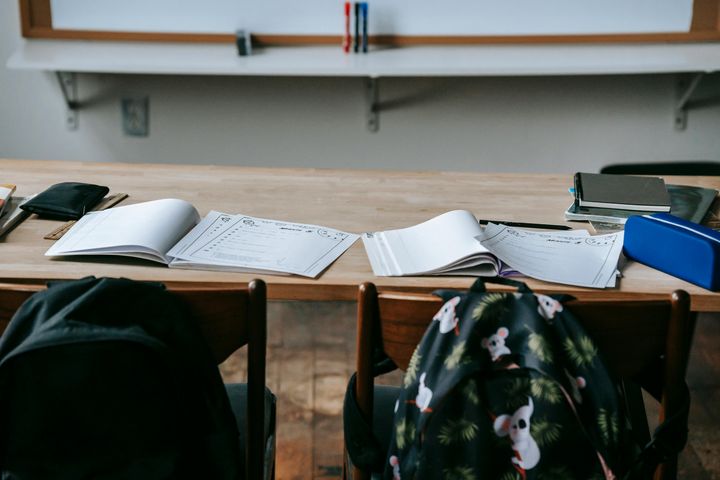 To skolepulter med åpne skrivebøker, arbeidsark og skolesekker hengende på stolene, sett forfra i et klasserom.