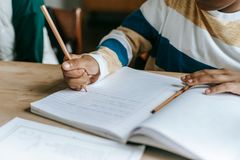 Et barn sitter ved en pult og skriver med blyant i en linjert skrivebok, med en åpen bok ved siden av.