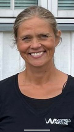 Portrett av en smilende kvinne med lyst hår i hestehale. Hun har på seg en svart t-skjorte med Vestfold museene -logo, og står foran en hvit trebygning.