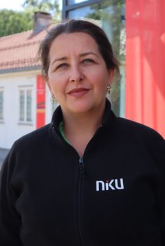 Portrett av en smilende kvinne med mørkt hår i hestehale. Hun har på en svart fleecejakke med NIKU-logo, og står foran en hvit trebygning.
