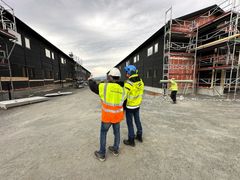 To personer iført verneutstyr og refleksvester står på en byggeplass og peker mot nye bygninger under oppføring.