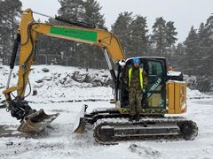 Mann med refleksvest står på beltet under førerhuset til gravemaskin.