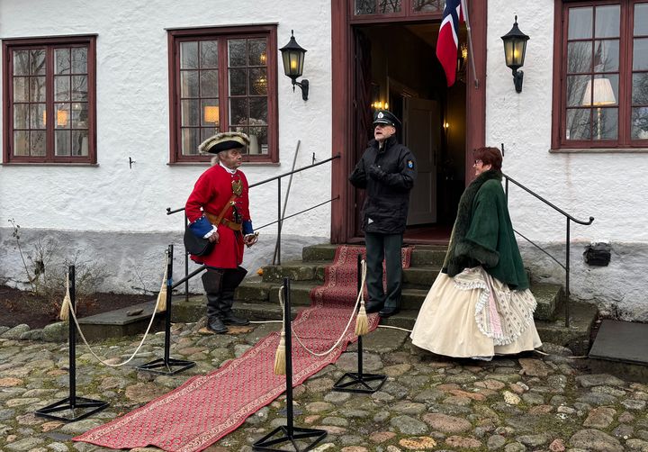 Mann i militær uniform taler på trapp med løper foran gammelt, hvitt hus med røde karmer. På hver side av han står henholdsvis en kvinne og en mann kledd i klær fra 1700-tallet.