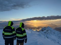 To personer i med ryggen til fotografen står på toppen av fjell og ser ut over horisonten i solnedgang.