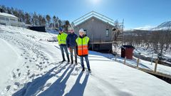 Tre personer i arbeidsklær foran et hus under bygging i et snødekt landskap.