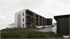 En moderne boligblokk med tre etasjer, sett fra veien. Spiraltrapp på byggets side, parkeringsplasser med biler foran. Utsikt mot sjøen og fjell i bakgrunnen.