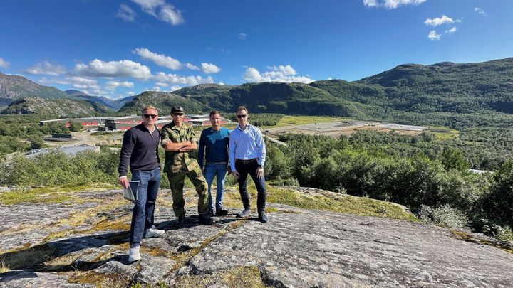 Fire menn står på en berghøyde med et stort anleggs og grønt område i bakgrunnen.