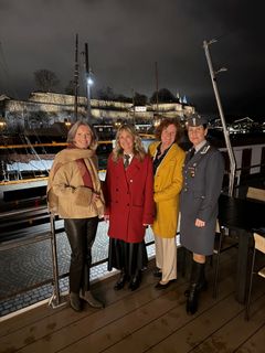 Fire kvinner står sammen utendørs på en brygge med en festning opplyst i bakgrunnen. En av dem er i militæruniform.
