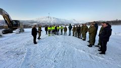 En gruppe mennesker står i snøen i en sirkel i et vinterlandskap med fjell i bakgrunnen. En stor gravemaskin er synlig til venstre.