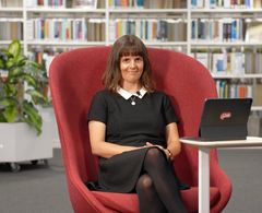 Kvinne i svart kjole sitter i rød stol med en bærbar datamaskin i et bibliotek.