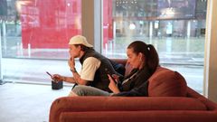 To personer sitter på en sofa og ser på telefonene sine.