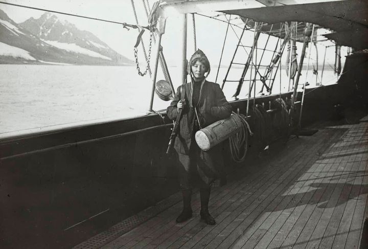 Hanna Resvoll-Holmsen (den gang Resvoll-Dieset) om bord på skipet ≪Princesse Alice≫, med botaniserkasse og gevar. Fotografiet er fra den første ekspedisjonen hun deltok i på Svalbard, i 1907. Fotografen er Gunnar Holmsen, som hun senere giftet seg med. Utsnitt av svart-hvitt-positiv på glass, lysbilde, 8 x 8 cm