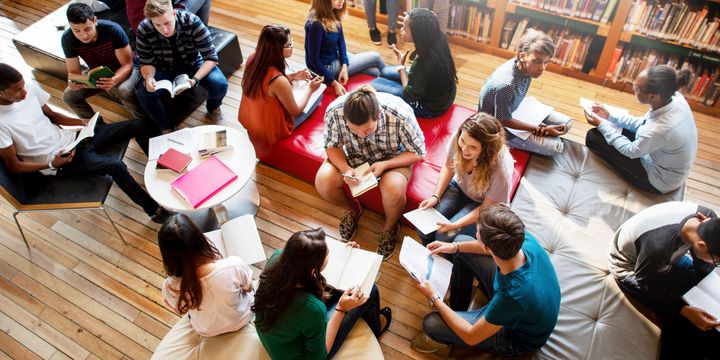 En gruppe unge voksne sitter i en sirkel på sofaer og gulv i et bibliotek, og de leser eller diskuterer.