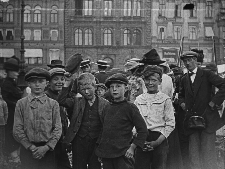 Fem%20unge%20gutter%20st%E5r%20foran%20en%20gruppe%20mennesker%20p%E5%20en%20travel%20gate%20i%20en%20by.%20Bakgrunnen%20viser%20flere%20bygninger.