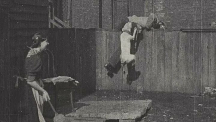 En svart-hvitt scene fra filmen "The Tramp and the Dog" (1896), som viser en person i en dramatisk situasjon sammen med en hund, mens en annen person ser på.