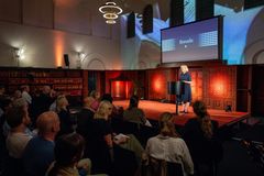 Alt text: Person står på scenen og holder en presentasjon foran publikum i en bibliotekssal.