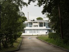 En smal vei med grønne trær og gress på hver side fører fram til en hvit bygning med store vinduer i turkis innramming.