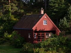 Rødmalt kvadratisk hytte med saltak og sprosser på vinduene. Hytta er plassert i tett gran- og furuskog.