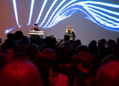 To unge personer opptrer på en scene, med lysshow på veggen i bakgrunnen. Foran dem står mange mennesker trengt sammen.