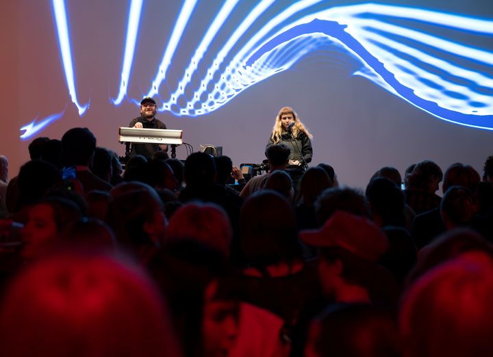 To%20unge%20personer%20opptrer%20p%E5%20en%20scene%2C%20med%20lysshow%20p%E5%20veggen%20i%20bakgrunnen.%20Foran%20dem%20st%E5r%20mange%20mennesker%20trengt%20sammen.