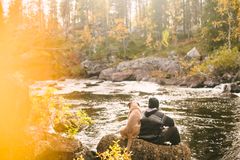 En person sitter ved en elv i skogen med en hund på en solfylt høstdag.