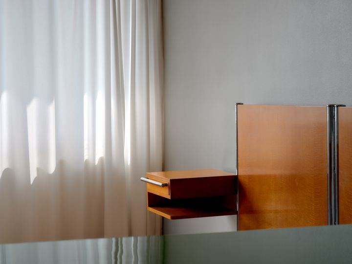 A%20white%20curtain%20through%20with%20light%20seeping%20in%2C%20sections%20of%20a%20bedside%20table%20and%20the%20headboard%20of%20a%20bed.