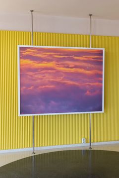 A photograph of a sunrise tinging clouds in the sky yellow, pink and purple, placed in front of on a bright yellow wall.