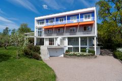 Facade of a rectangular brick villa with flat roof and large windows. White with blue details and orange awnings partly extended.