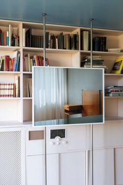 A photograph of a white curtain through with light seeping in, sections of a bedside table and the headboard of a bed placed in front of a bookshelf.