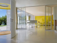 Music room with grand piano and simple furnishings, one yellow wall, otherwise all white. Open door to the garden, giving a glimpse of a sculpture and green plants.
