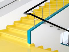 White stairwell in a sanatorium, with yellow steps and black handrail on metal spindles fixed to turquoise base.