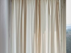White curtain pulled slightly to one side to give a glimpse of the sky and the landscape outside. Sunlight on the curtain.