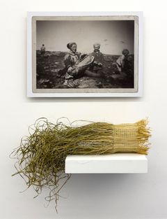 Two works, a photo outdoors showing a woman dressed in a Sami costume, two childen on her side and a man in the background. Underneath the photo a necklace made of a bundle of straw hold together by a silver thread.